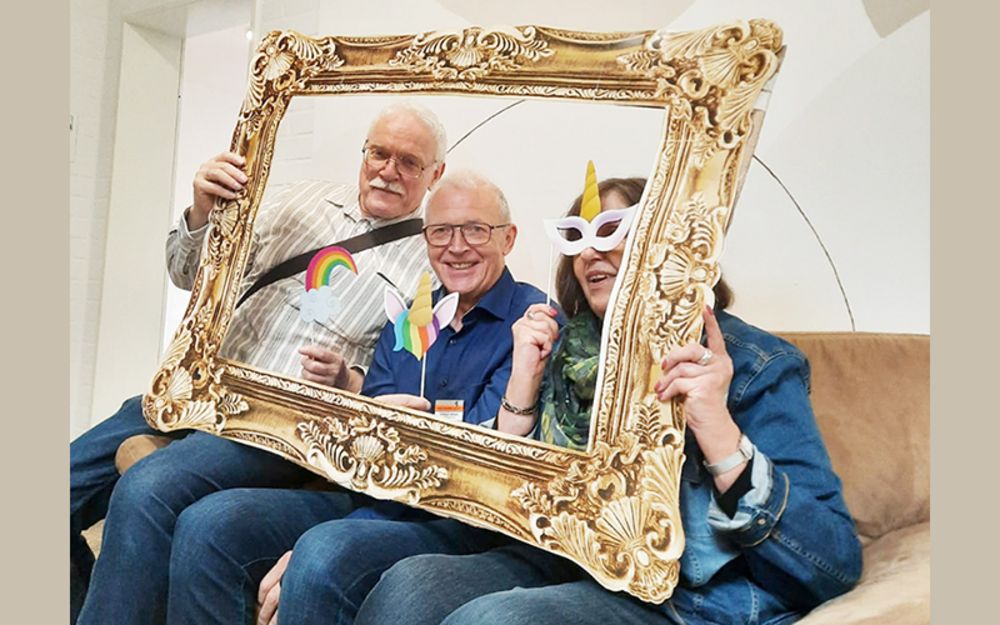 Foto zeigt drei Personen, auf einem Sofa sitzen und sich fotografieren lassen. Um sich herum halten sie einen Bilderrahmen hoch.