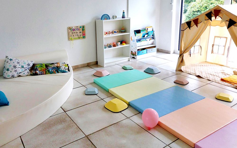 Foto zeigt den Sensomotorik-Raum. Er ist mit Matten in zarten Farben, einem Zelt, Regal mit Spielsachen, Büchern und vielen Kissen eingerichtet.