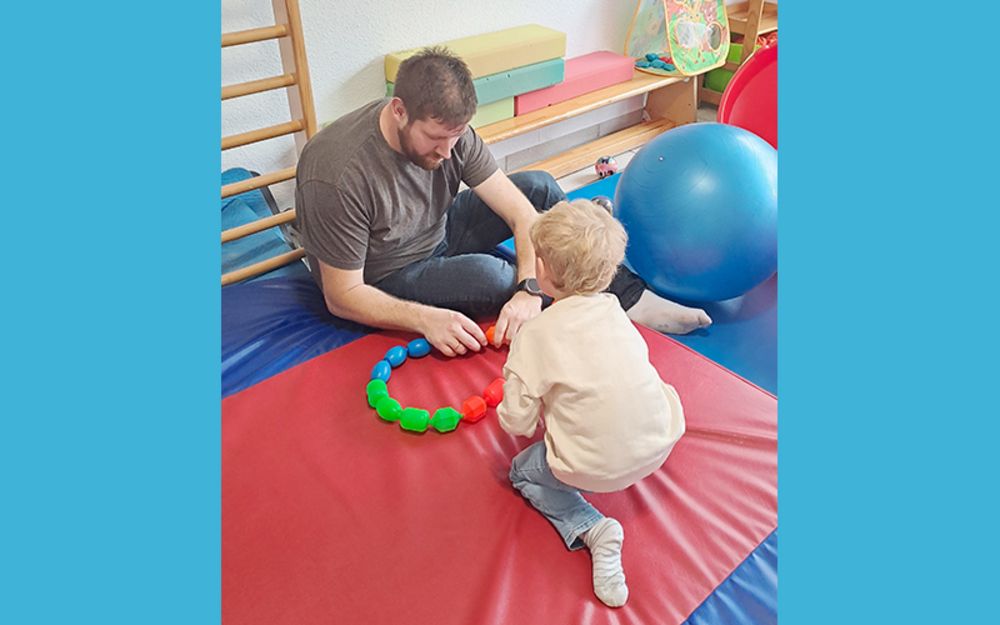 Foto zeigt einen Mann und ein Kleinkind, die in einem Raum spielen, in dem viele bunte Matten ausgelegt sind.