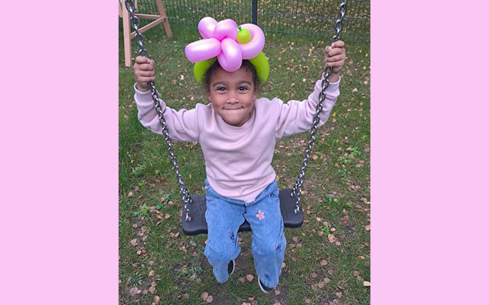 Foto zeigt ein Mädchen draußen auf der Schaukel. Sie hat ein bunte Blume aus Luftballons auf dem Kopf.