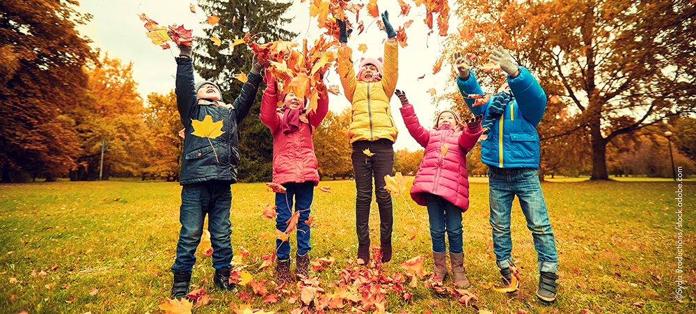 Foto zeigt Kinder auf einer Rasenfläche, die Herbstlaub in die Luft werfen